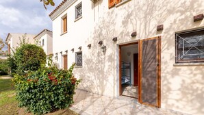 A white building with a tiled entrance, a wooden door, and a small garden with red flowers.