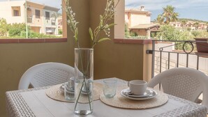 A balcony with a table set for two, featuring a clear vase with a plant, two glasses, and a cup. The balcony overlooks a residential area with houses and palm trees.