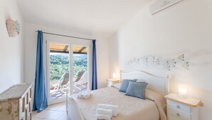 A bedroom with a bed, bedside tables, a window with curtains, and a view of greenery outside. - Three-room apartment with 6 beds, sea view, about 65/70 sq m, living room with kitchenette, double or twin sofa bed, double bedroom, one twin bedded room or with bunk bed, bathroom. Side or front sea view. <br>Apartments arranged on different levels. Spa (Baja Sardinia)
