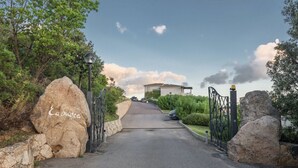 A gated entrance with large rocks and a lamp post, leading to a building with a white facade and a red roof. - Three-room apartment with 6 beds, sea view, about 65/70 sq m, living room with kitchenette, double or twin sofa bed, double bedroom, one twin bedded room or with bunk bed, bathroom. Side or front sea view. <br>Apartments arranged on different levels. Spa (Baja Sardinia)