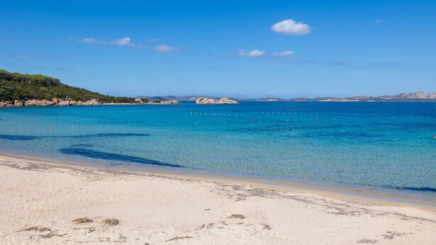 A beach with clear turquoise water, sandy shore, and distant rocky formations. - Three-room apartment with 6 beds, sea view, about 65/70 sq m, living room with kitchenette, double or twin sofa bed, double bedroom, one twin bedded room or with bunk bed, bathroom. Side or front sea view. <br>Apartments arranged on different levels. Spa (Baja Sardinia)