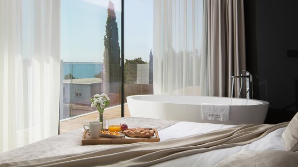 A bedroom with a large window, a bathtub, and a breakfast tray on the bed.