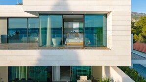 A modern two-story house with large glass windows, a balcony with a view of the sea, and a bedroom with a bed and a bathtub.