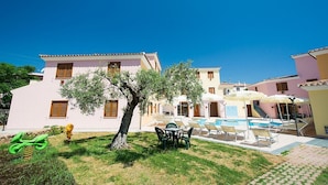 A building with a pool, outdoor seating, and a tree. - Two-room apartment with 4 beds, about 38 sq m, living room with kitchenette, sofa bed or bunk bed, bedroom with double bed (separable beds), bathroom with shower. Possibility 1 infant in cot. <br>The apartments are arranged over three floors (ground, fir (Sos Alinos)
