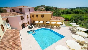 A swimming pool surrounded by lounge chairs and umbrellas, with buildings in the background. - Two-room apartment with 4 beds, about 38 sq m, living room with kitchenette, sofa bed or bunk bed, bedroom with double bed (separable beds), bathroom with shower. Possibility 1 infant in cot. <br>The apartments are arranged over three floors (ground, fir (Sos Alinos)
