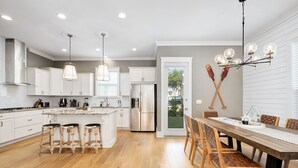 A modern kitchen with white cabinets, a central island with stools, stainless steel appliances, and a dining area with wooden chairs. - Amazing Beach house Steps from 30a | Golf Cart | Beach, Pool & Lake access! (Inlet Beach)