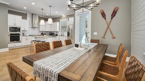 A modern kitchen with white cabinets, a wooden dining table, and a chandelier. - Amazing Beach house Steps from 30a | Golf Cart | Beach, Pool & Lake access! (Inlet Beach)