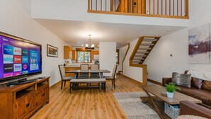 A spacious living area with a wooden floor, a dining table with chairs, a television on a wooden stand, a staircase, and a kitchen in the background. - Rendezvous at Northstar_3 Story High End Condo, Nest, Alexa, Remote Fireplace, Smart TV's +NPOA Pass (Truckee)