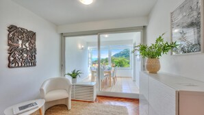 Ein modernes Wohnzimmer mit einem weißen Sofa, einem kleinen Tisch, einer Wanddekoration, einem Blumentopf und einer Glastür, die auf einen Balkon mit Blick auf Grün führt.