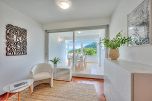 A modern living room with a white sofa, a small table, a decorative wall piece, a potted plant, and a sliding glass door leading to a balcony with a view of greenery. - Residenza Parco Carona (Lugano)