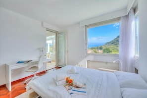 A hotel room with a bed, a desk, a chair, a window with a view of a mountain, and a door leading to an outdoor area. - Residenza Parco Carona (Lugano)