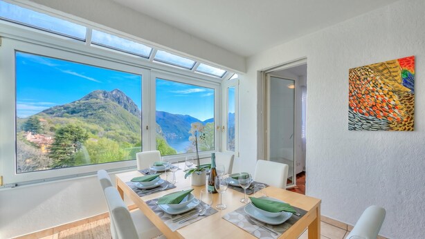 Ein Essbereich mit Tisch für vier Personen, großen Fenstern mit Blick auf die Berge und einem farbenfrohen, abstrakten Gemälde an der Wand.