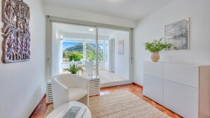 Ein modernes Wohnzimmer mit einem weißen Sofa, einem kleinen Tisch, einem Schrank, einem großen Fenster und Blick auf einen Balkon mit Gartenmöbeln.