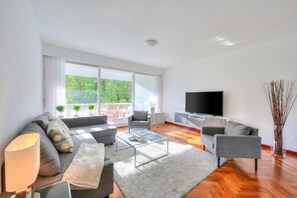 A modern living room with a sofa, armchair, coffee table, TV, and a large window with a view of greenery. - Residenza Parco Carona (Lugano)