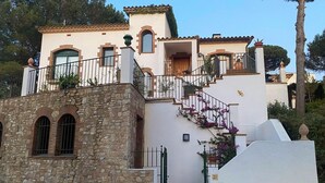 Un edifici de diverses plantes amb façana de pedra, una escala amb barana de ferro forjat i un balcó amb plantes en test.