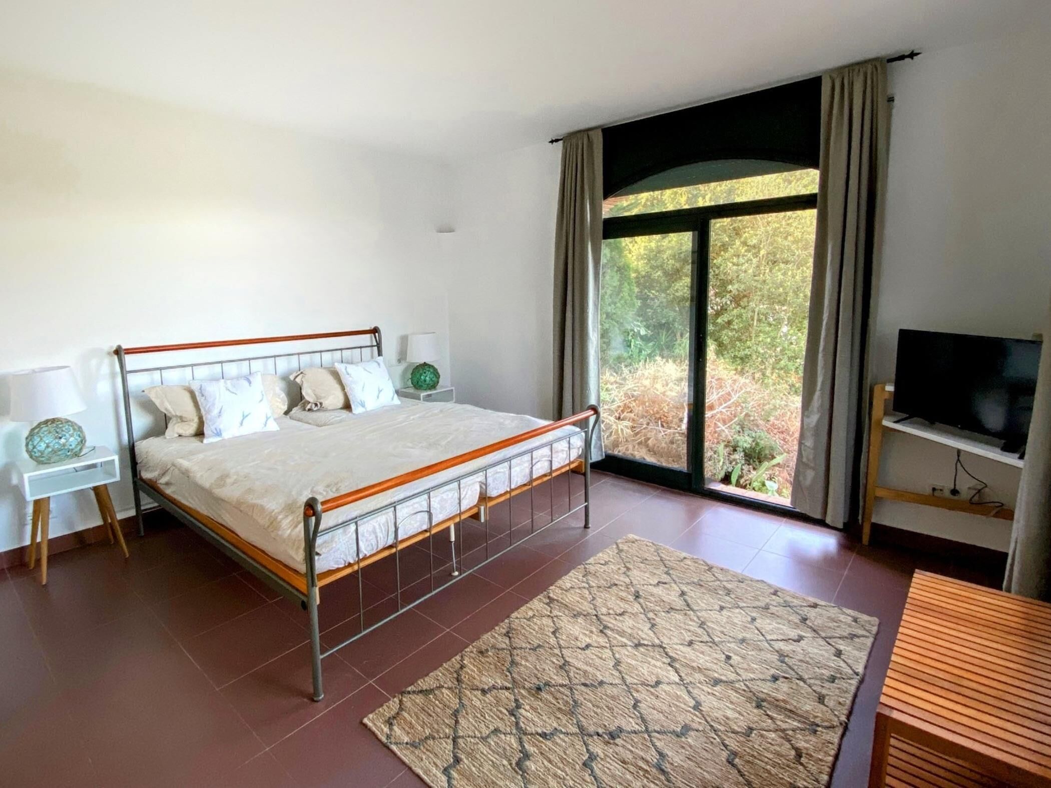 A bedroom with a bed, bedside tables, a TV, and a view of the outdoors.