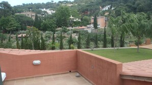 Um terraço com chão de azulejos, luminária na parede e vista para uma paisagem verde com árvores e edifícios ao fundo.