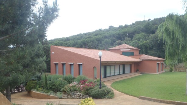 Um edifício com telhado de telhas vermelhas, entrada por um caminho de paralelepípedos e gramado bem cuidado.