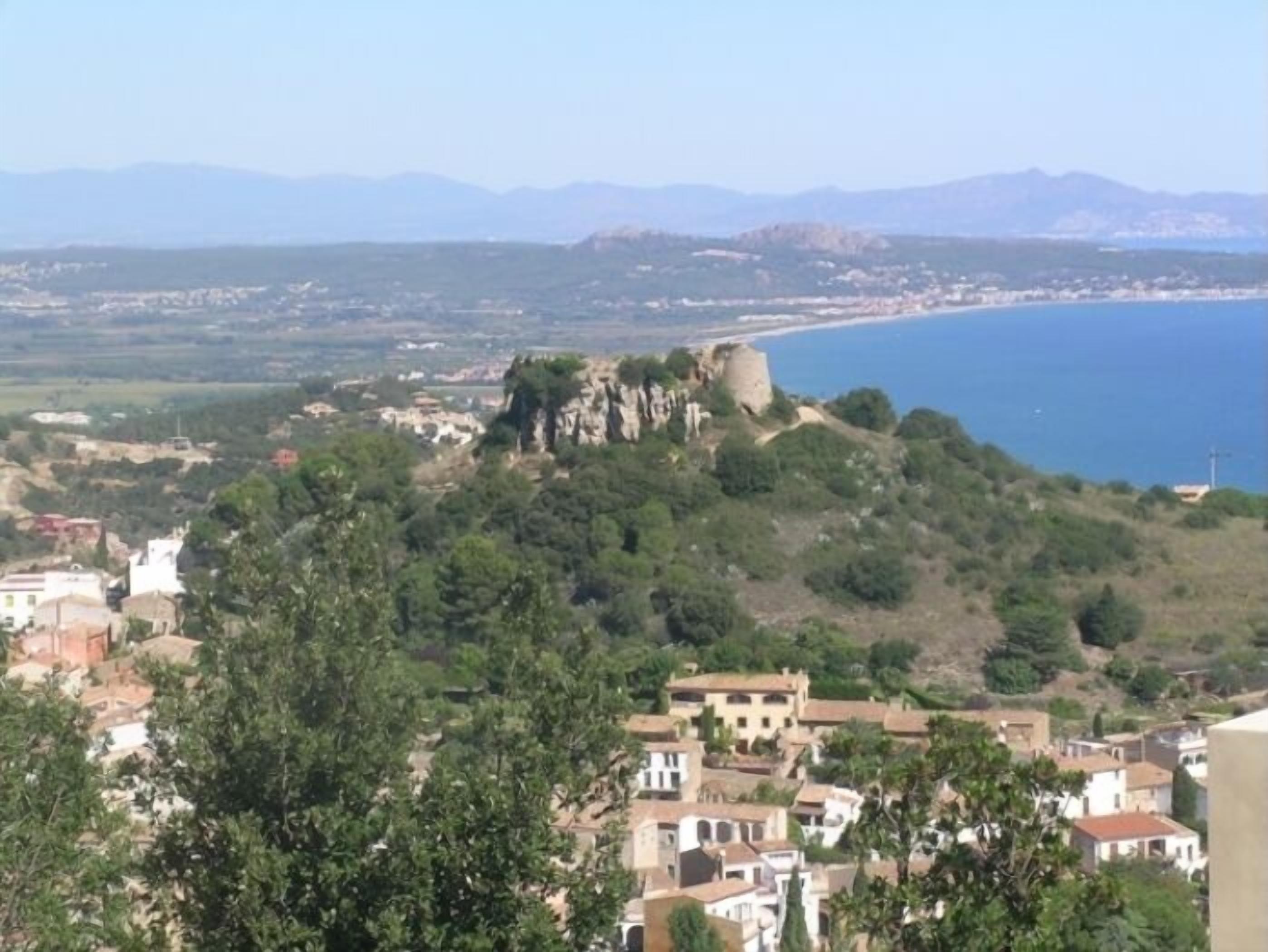 Un pueblo costero con un castillo imponente en una colina, rodeado de vegetación y edificios.