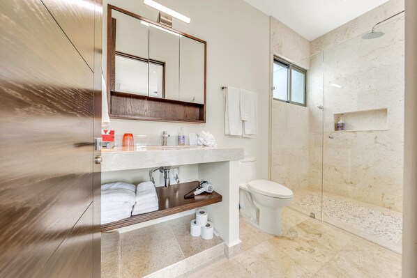 Un baño moderno con ducha de mármol, un espejo grande y tocador con artículos de aseo.