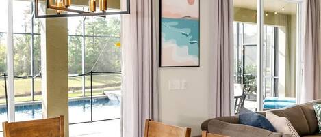 A dining area with a wooden table set for four, surrounded by chairs, with a view of a swimming pool and a sunset painting on the wall.