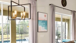 A dining area with a wooden table set for four, surrounded by chairs. A large window with curtains allows natural light to enter. - Southwest facing pool! 5 Miles from Disney. (Kissimmee)