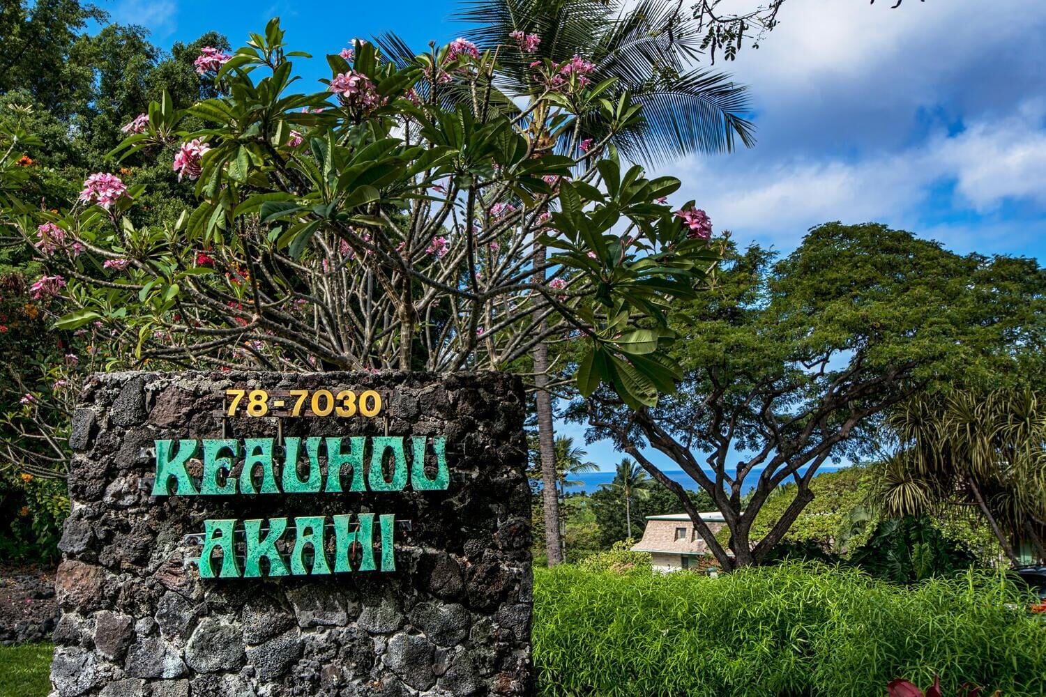 A stone sign with the number "78-7030" and the name "Kauhou Akani" in a tropical setting with lush greenery and a clear sky.