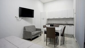 Un espacio habitable compacto con sofá, una mesa de comedor con sillas, una zona de cocina con armarios y un televisor de pantalla plana montado en la pared.