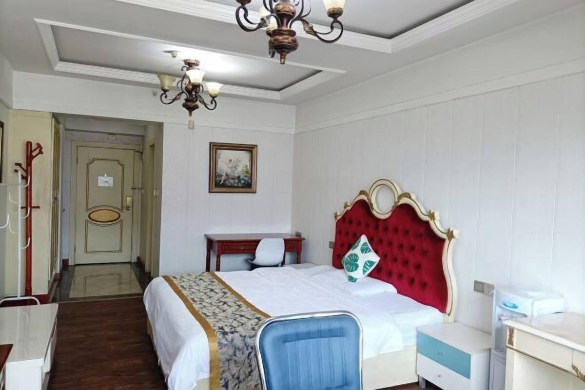 A hotel room with a bed, a desk, a chair, a nightstand, and a chandelier.