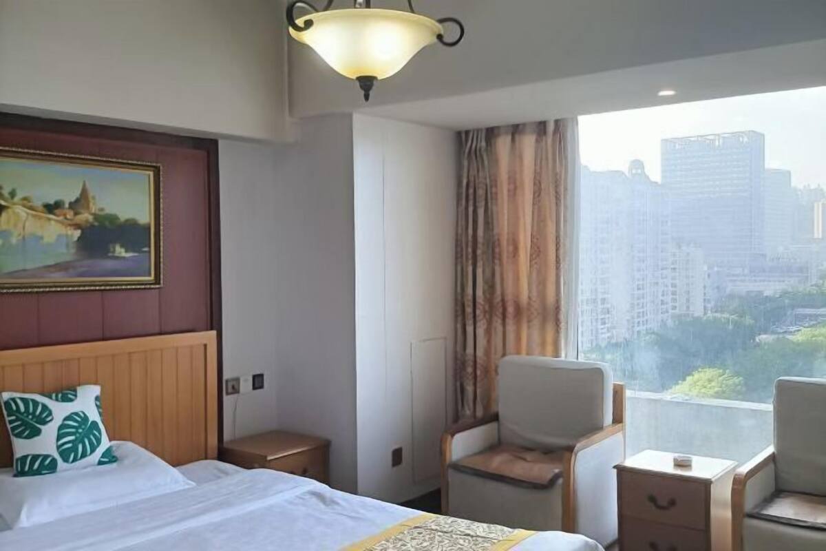 A hotel room with a bed, a chair, a nightstand, a painting, and a window with a city view.