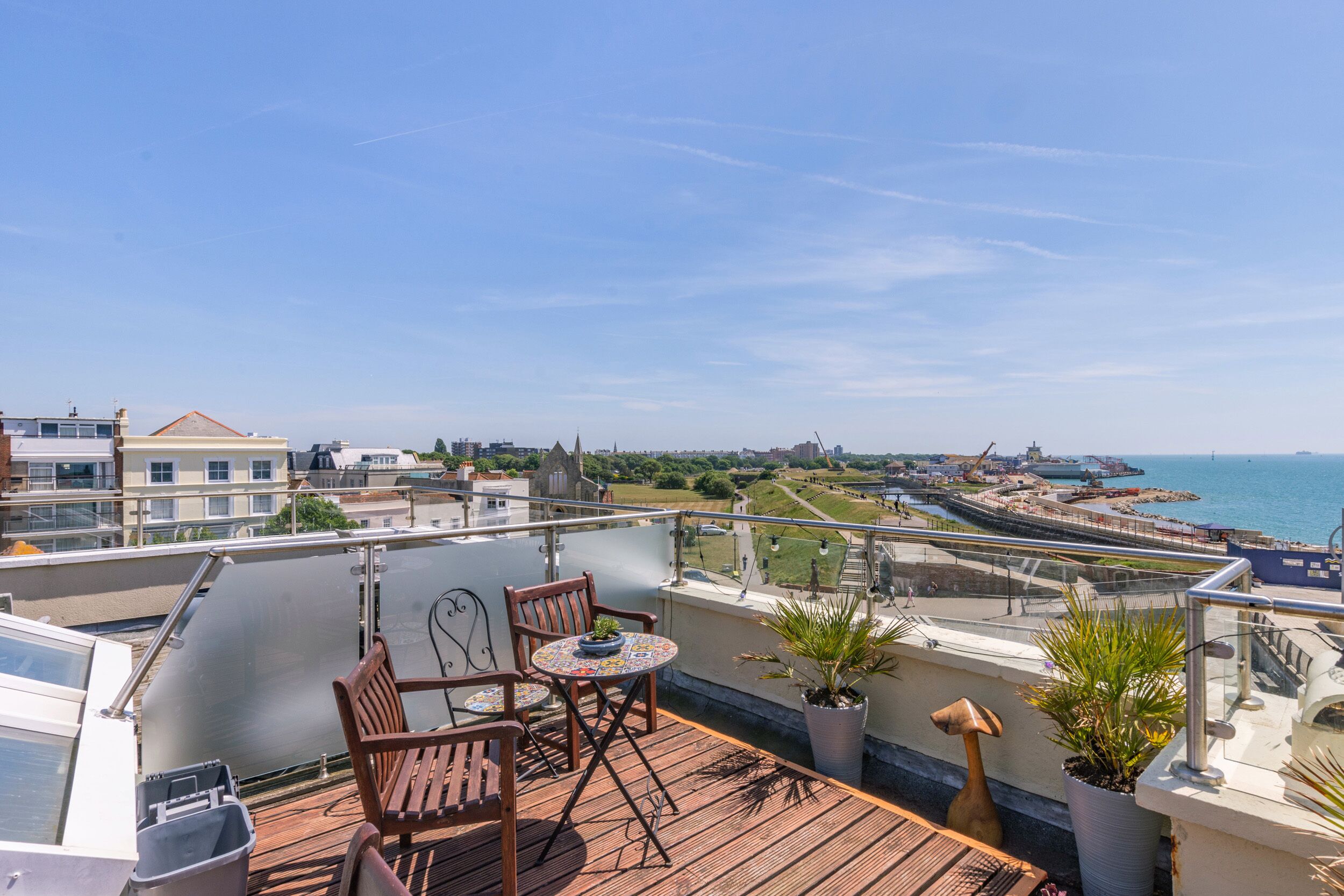A balcony with a wooden table and chairs, potted plants, and a view of the sea and cityscape.