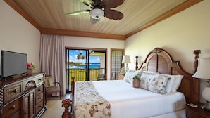 A hotel room with a large bed, wooden headboard, a ceiling fan, a television, a desk with a lamp, and a view of the ocean through a sliding door. - Poipu Sands 522 Gorgeous Oceanfront  By Suite Paradise (Koloa)
