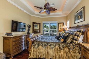 A bedroom with a large bed, a ceiling fan, a TV, a dresser, and a window with a view of greenery.