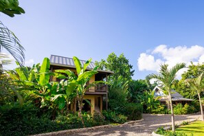 Ein zweistöckiges Haus mit Balkon, umgeben von üppiger Vegetation und blauem Himmel.