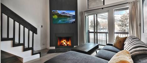 A modern living room with a fireplace, a large TV, and a sofa with a view of the outdoors.