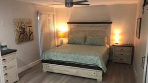 Camera da letto con un letto, lampade da comodino, ventilatore a soffitto, comodino, cassettiera e un quadro appeso al muro.