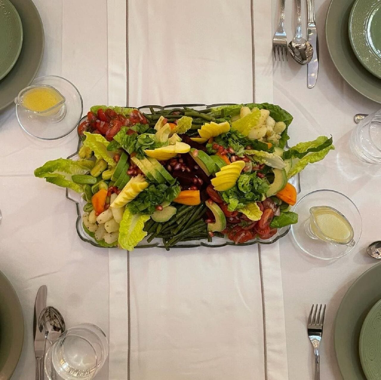 Mesa preparada para una comida con una ensalada grande como centro de mesa, recipientes de vidrio transparente para condimentos y cubiertos.