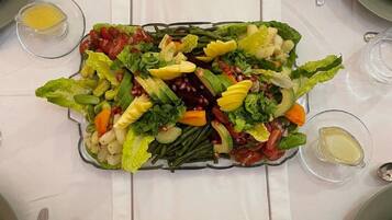 Mesa preparada para una comida con una ensalada grande como centro de mesa, recipientes de vidrio transparente para condimentos y cubiertos.