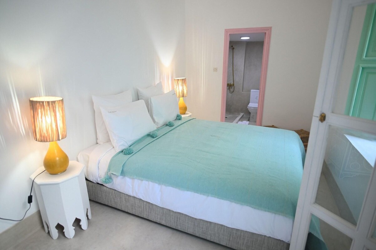 A bedroom with a bed, two bedside lamps, a green blanket, and a door leading to a bathroom.