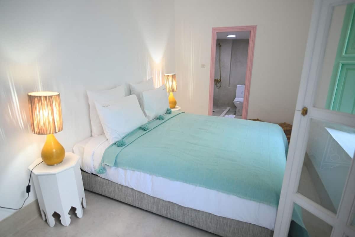 Un dormitorio con una cama, dos lámparas de noche, una manta verde y una puerta que da al baño.