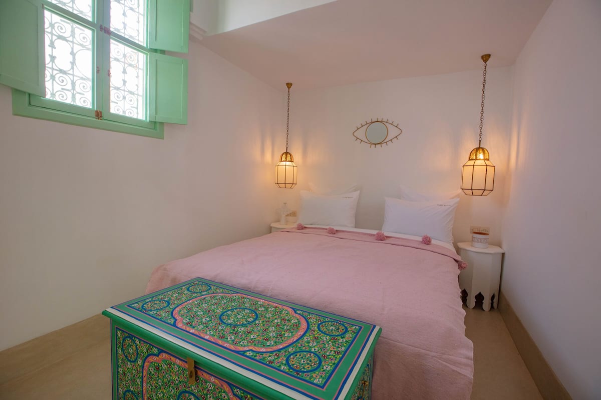 Un dormitorio con cama, mesitas de noche, un cofre decorativo y una ventana con contraventanas.