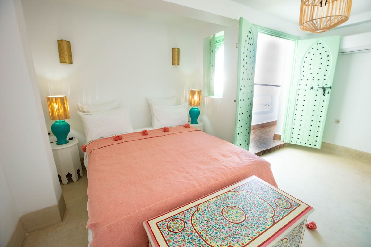 Un dormitorio con una cama, dos mesitas de noche con lámparas, una pequeña mesa con mantel estampado y una puerta verde.