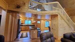 Un intérieur de chalet en bois spacieux, comprenant une cuisine, un coin repas et une chambre à l’étage, accessible par un escalier en bois.