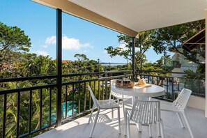Un balcon avec une table ronde et des chaises, donnant sur une piscine et une végétation luxuriante.