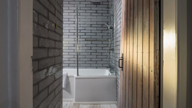 A bathroom with a brick wall, a white bathtub, and a wooden door. - Esmor Cottage (Caerwys)