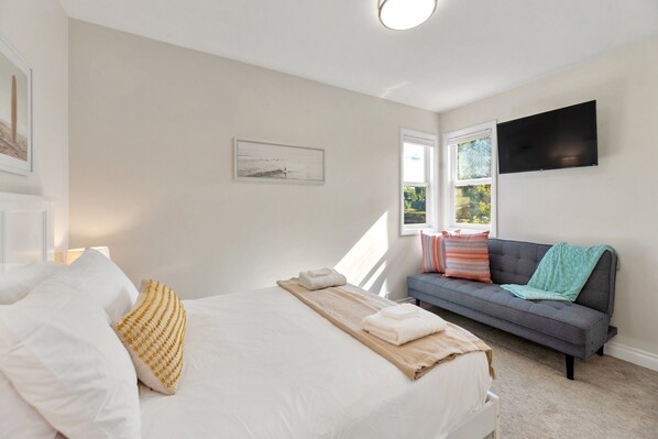 A bedroom with a bed, a gray sofa, a flat-screen TV mounted on the wall, and a window providing natural light.