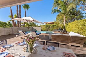 An outdoor pool area with lounge chairs, a dining table set for four, and a large umbrella.