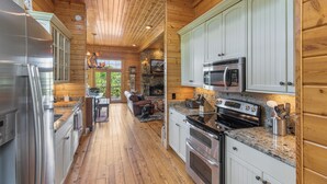 A log cabin interior with a kitchen featuring stainless steel appliances, white cabinets, and a stone fireplace.