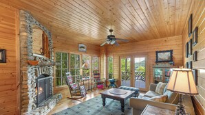 A cozy log cabin interior with a stone fireplace, wooden ceiling, and a comfortable seating area with a sofa and armchairs.