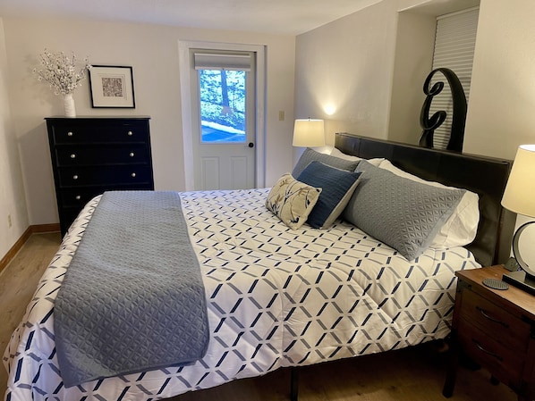 Un dormitorio con cama, cómoda, mesita de noche, lámpara, cuadro enmarcado y puerta que da a vistas de árboles.